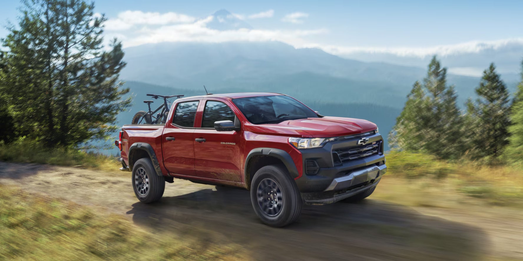 A red Chevrolet Colorado driving on a dirt road by a cliffside with a bike in its truck bed.