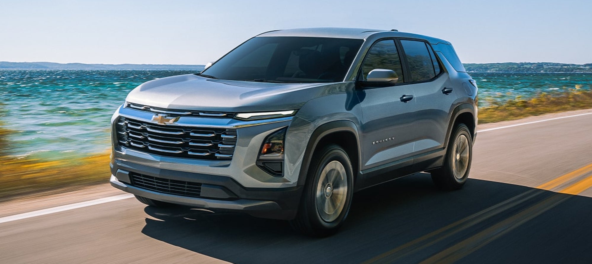 A silver Chevy Equinox driving down the road by the ocean.