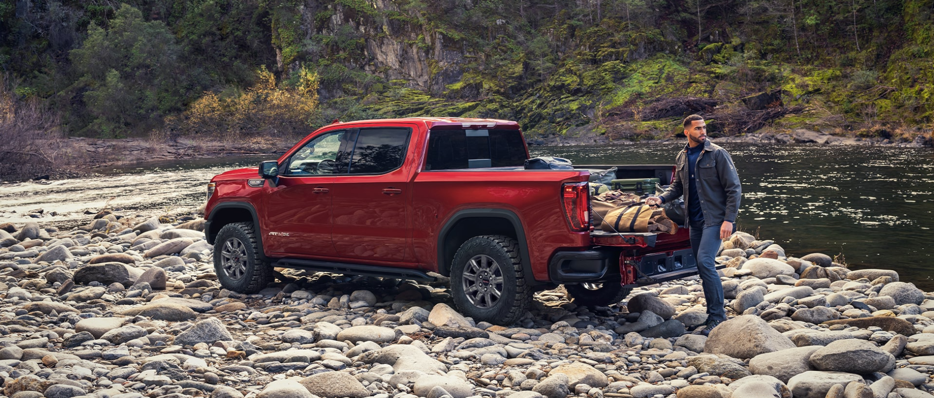 A red GMC Sierra 1500 off-road by a lake with a man loading up the truck bed.